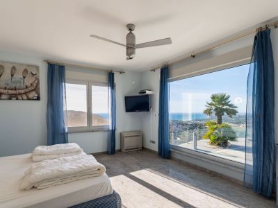 Helles Schlafzimmer mit großem Panoramafenster, direktem Meerblick und Palme. Heller Granitfliesenboden und blaue transparente Vorhänge.