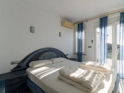 Schlafzimmer mit blau-dunklem Doppelbett und passenden Nachttischen. Glastüren mit blauen Vorhängen führen zur Terrasse, ergänzt durch eine Klimaanlage.