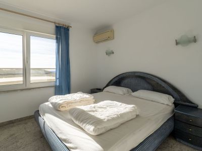 Schlafzimmer mit blau gemustertem Bett und geflochtenem Kopfteil, hellen Fliesenboden und Außenblick durch das Fenster. Eine Klimaanlage und passender Nachttisch sind vorhanden.