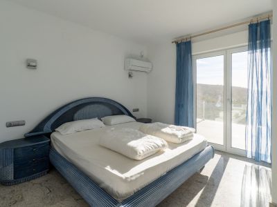 Schlafzimmer mit blauem Designbett und passenden Nachttischen, hellem Boden und direktem Terrassenzugang durch Glastür, mit Hügelaussicht und Tageslicht.