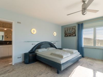 Schlafzimmer mit einem blau getönten Designerbett, passenden Nachttischen und hellem Marmorboden. Das Fenster bietet Hügelblick, ergänzt durch blaue Vorhänge und ein angrenzendes Badezimmer.