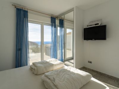 Schlafzimmer mit weiß bezogenem Bett, Wandfernseher und großen Glasschiebetüren mit blauen Gardinen, die Zugang zu einer Terrasse mit Panoramameerblick bieten.