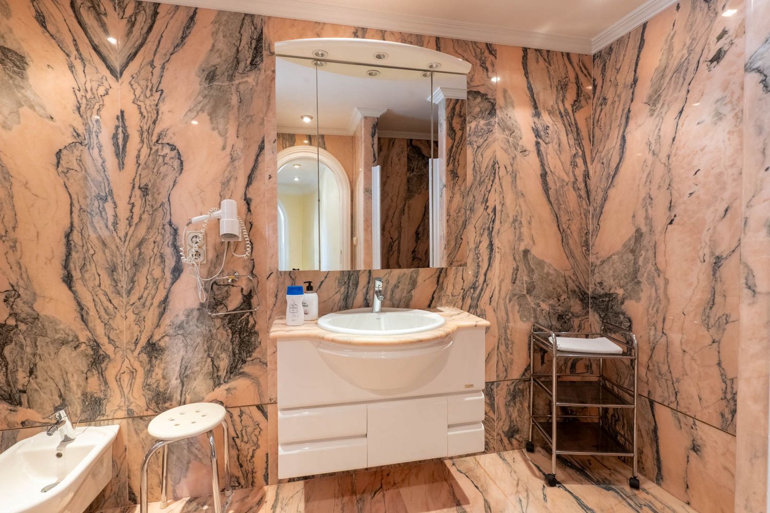 Badezimmer mit auffälligem rosa Marmor und dunklen Adern, ausgestattet mit einem schwebenden Waschtisch, beleuchtetem Spiegelschrank und Bidet.