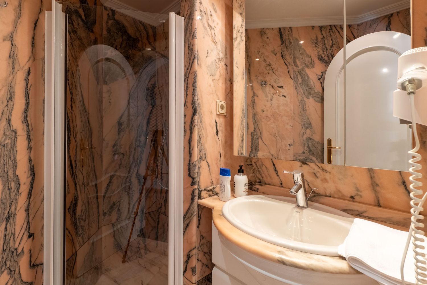 Badezimmer mit markanten rosa-schwarz geäderten Marmorwänden, einem weißen Keramikwaschbecken und einer Glasduschkabine mit Chromarmatur.