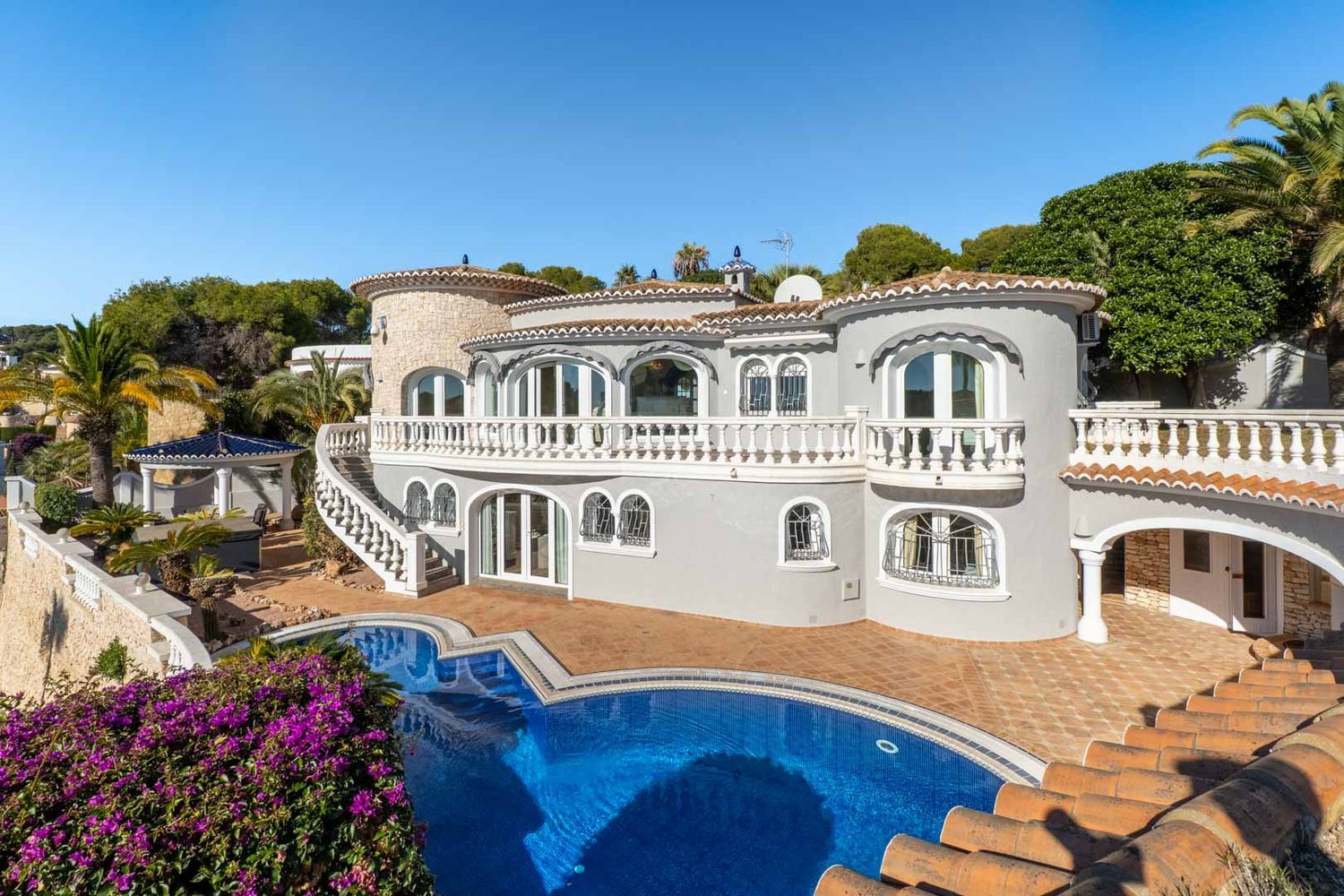 Die Villa zeigt eine graue Fassade, weiße Balustraden und einen markanten Privatpool. Eine geschwungene Treppe verbindet den oberen Balkon mit der terrakotta-gefliesten Poolterrasse, umgeben von mediterraner Flora.