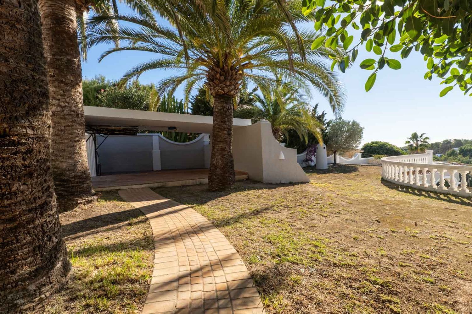 Ein gepflasterter Weg führt durch einen Garten mit Palmen und Rasen. Ein überdachter Carport und eine geschwungene weiße Balustrade sind sichtbar, umgeben von mediterraner Vegetation unter blauem Himmel.