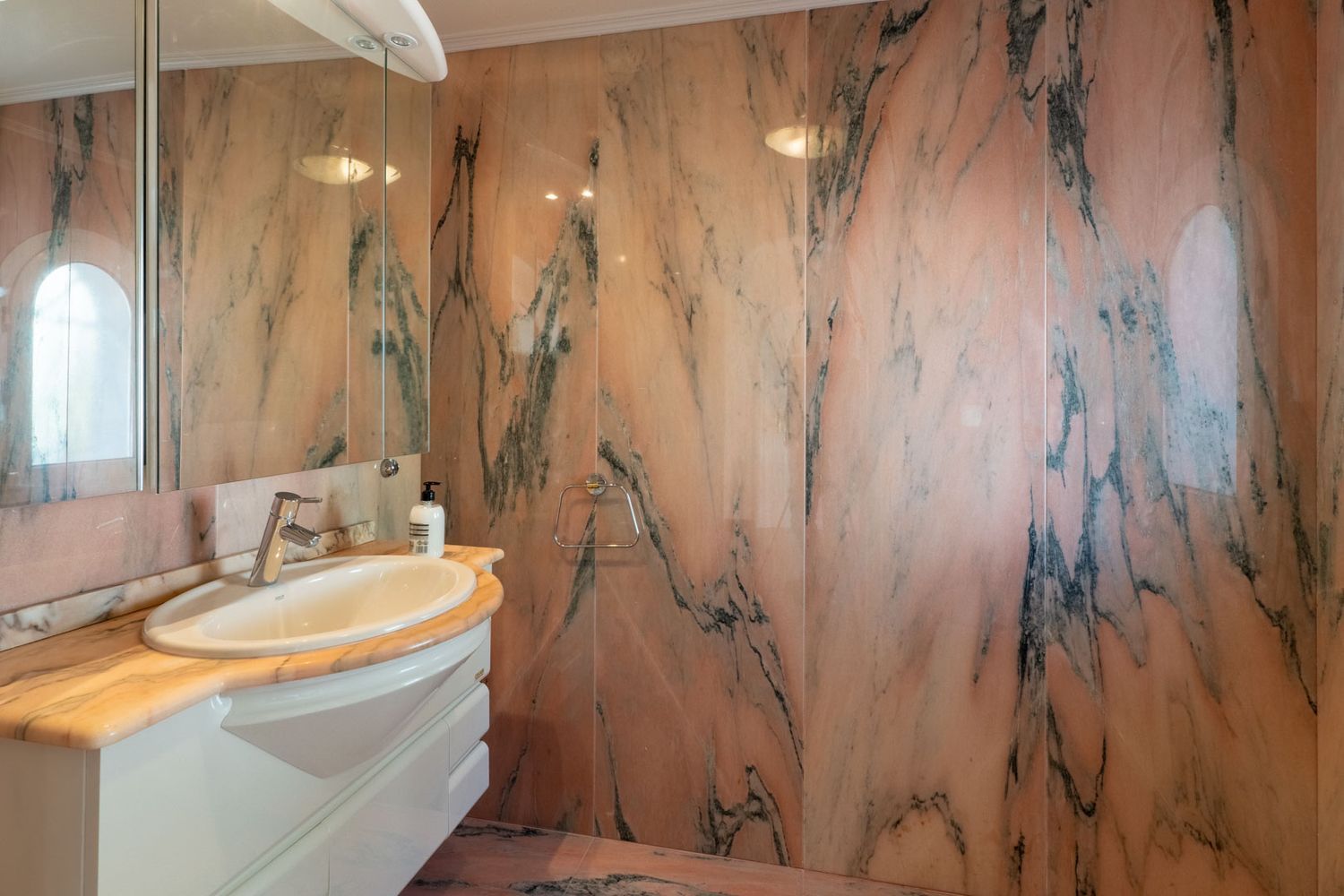 Elegantes Badezimmer mit Wänden und Waschtischplatte aus markantem rosa Marmor mit dunklen Adern. Ein weißer Waschtisch mit ovalem Becken und Spiegelschrank bietet Funktionalität.