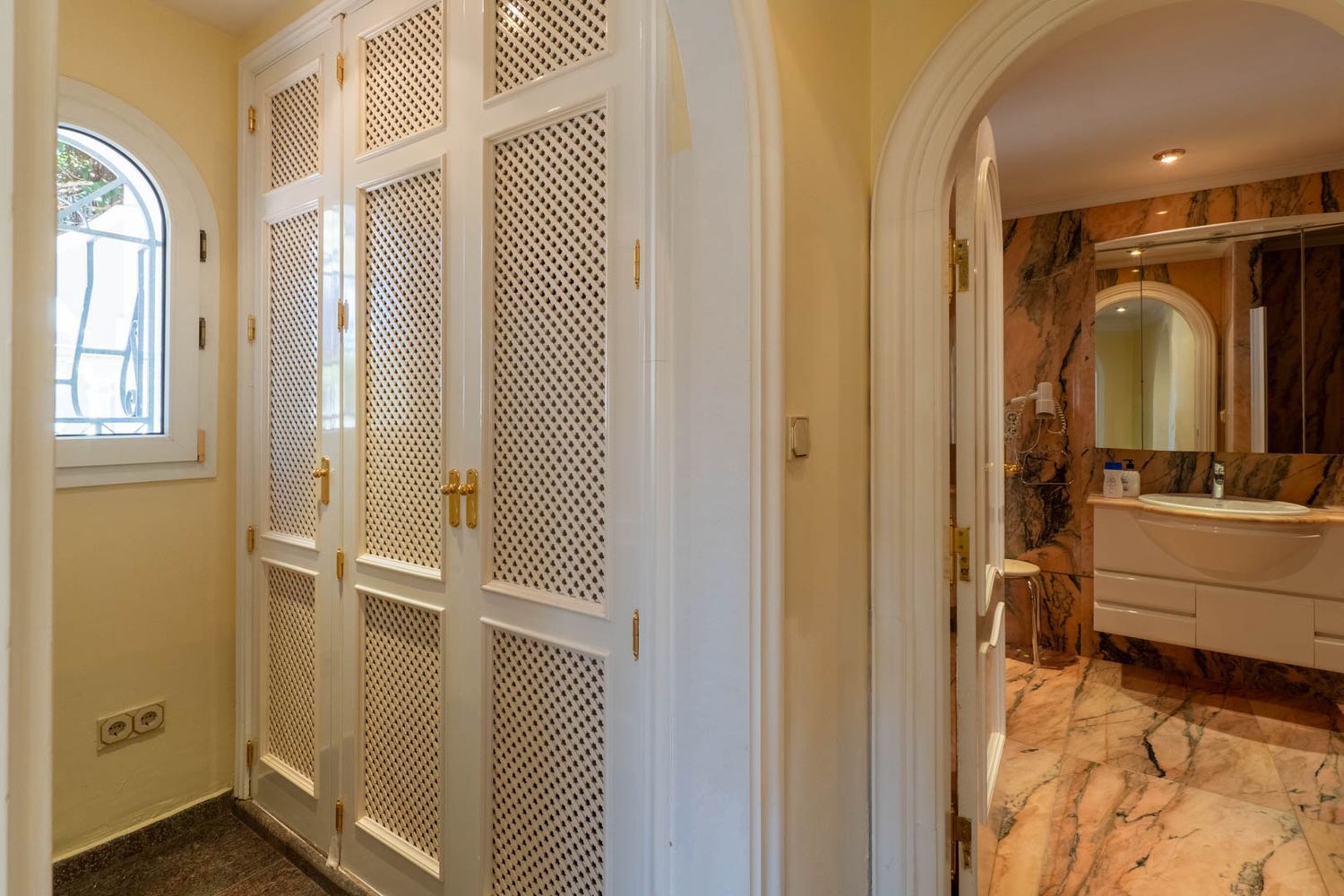 Flur mit weißen Einbauschränken mit Gittertüren und goldenen Griffen, neben einem Bogenfenster. Ein Rundbogen führt zu einem Badezimmer mit marmorierten Oberflächen.