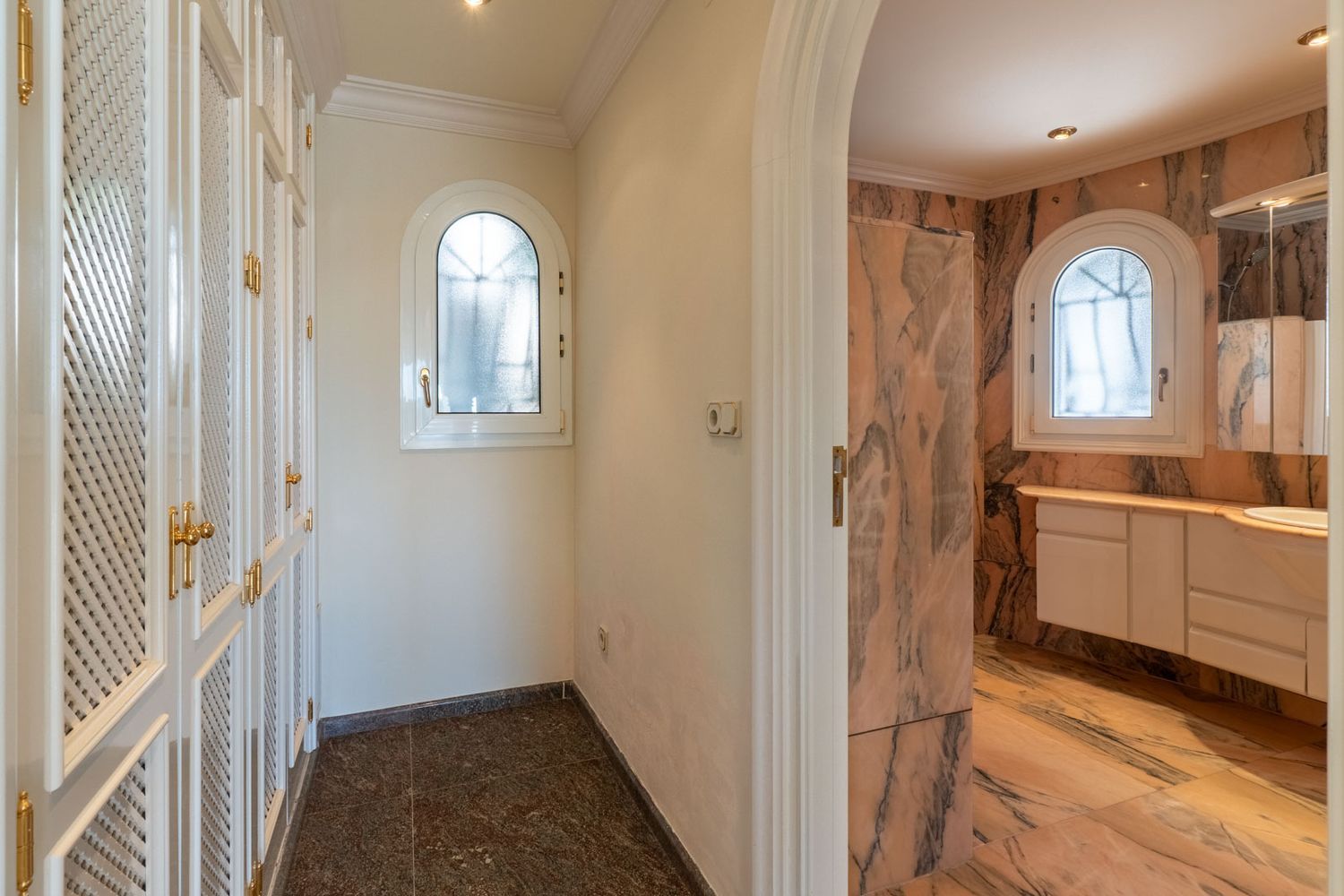 Flur mit weißen Lamellenschränken und goldenen Griffen, der zu einem Badezimmer mit rosa Marmorverkleidung und zwei Rundbogenfenstern führt.