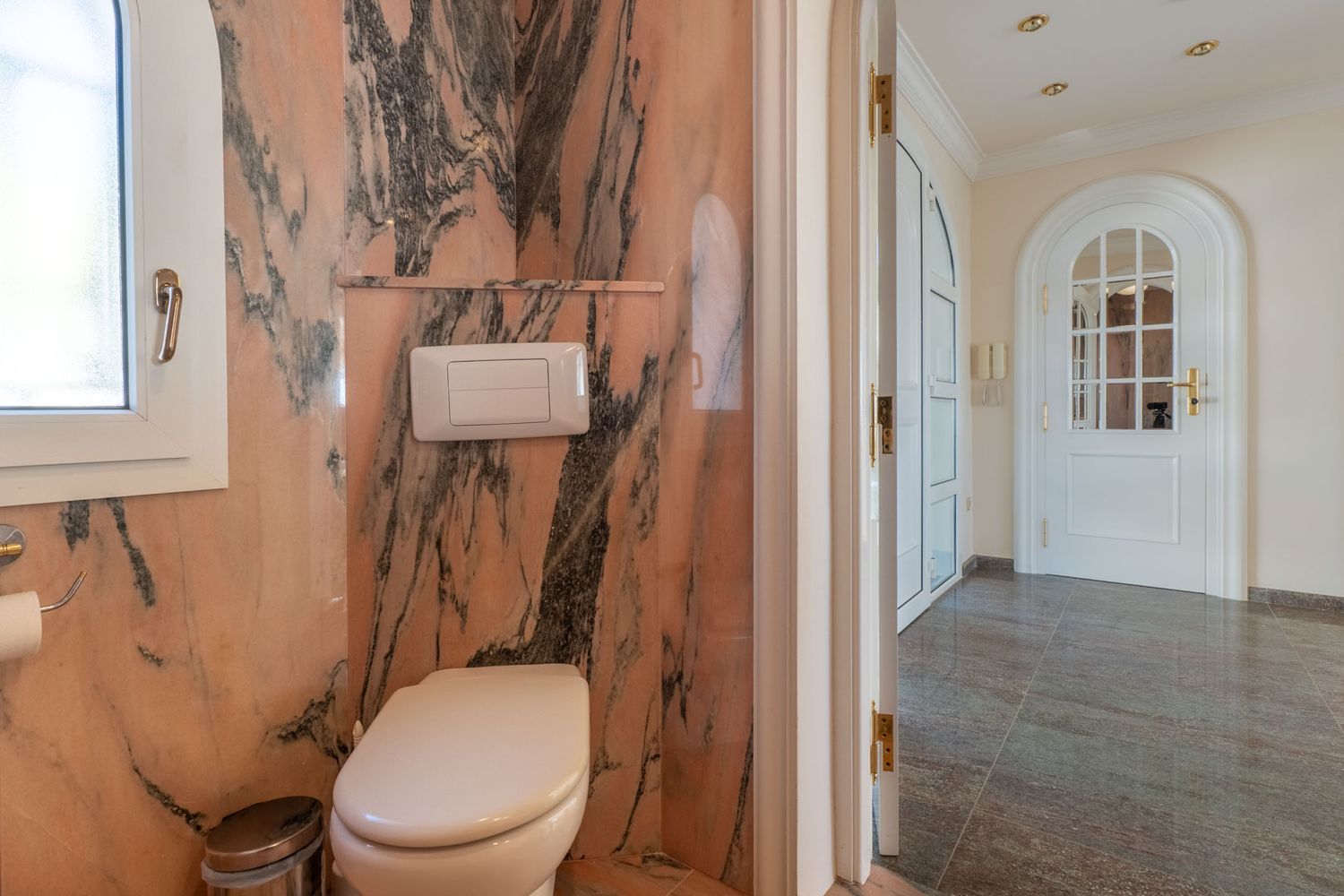 Gäste-WC mit rosa geäderten Marmorwänden und Fenster, das zu einem Flur mit dunklen Fliesen und einer Bogentür führt.