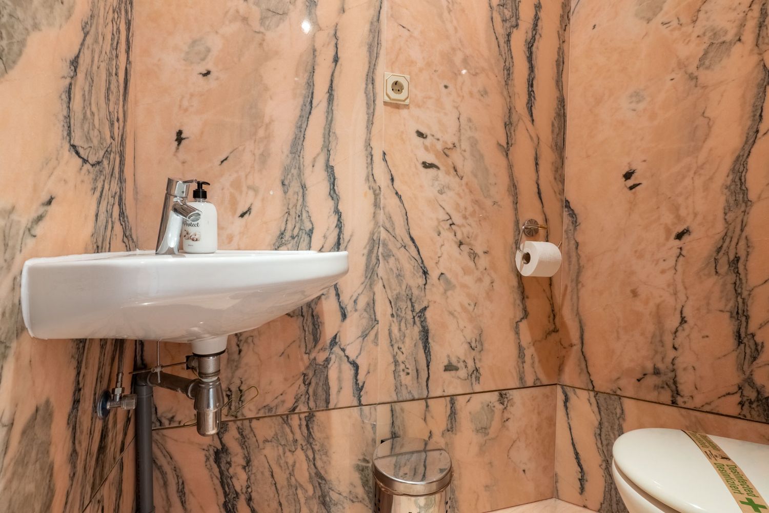 Gäste-WC mit Wänden und Boden aus rosa geädertem Marmor. Ausgestattet mit einem wandmontierten weißen Waschbecken, Chromarmatur und Toilette.