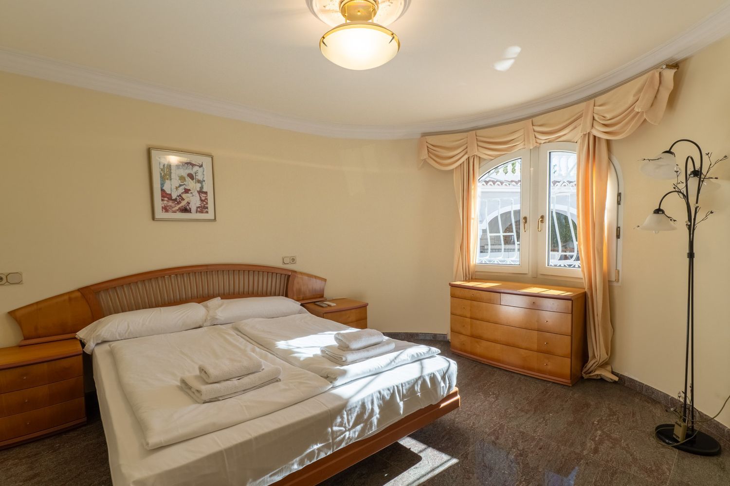 Geräumiges Schlafzimmer mit Doppelbett, Holz-Kopfteil und passenden Nachttischen. Natürliches Licht fällt durch ein geschwungenes Fenster auf den dunklen Fliesenboden.