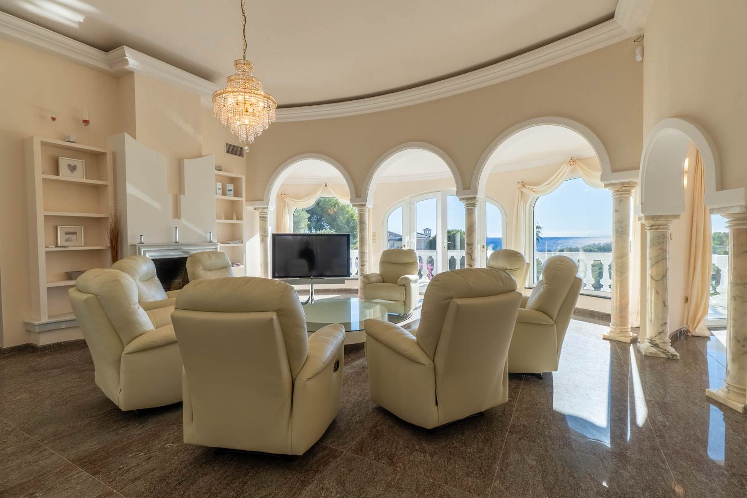 Geschwungenes Wohnzimmer mit Relaxsesseln, poliertem Marmorboden und Meerblick durch Bogenfenster. Ein zentraler Kristallleuchter und klassische Säulen prägen den eleganten Raum.