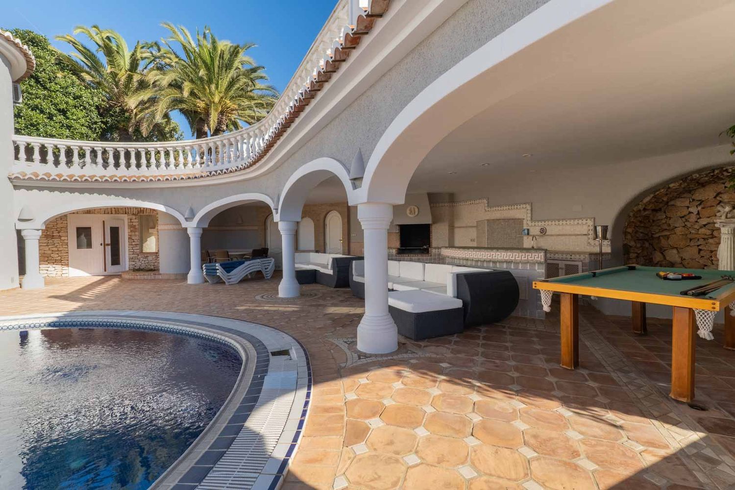 Große überdachte Terrasse mit geschwungenem Pool, Außenküche, Loungebereich und Billardtisch auf Terrakottafliesen.