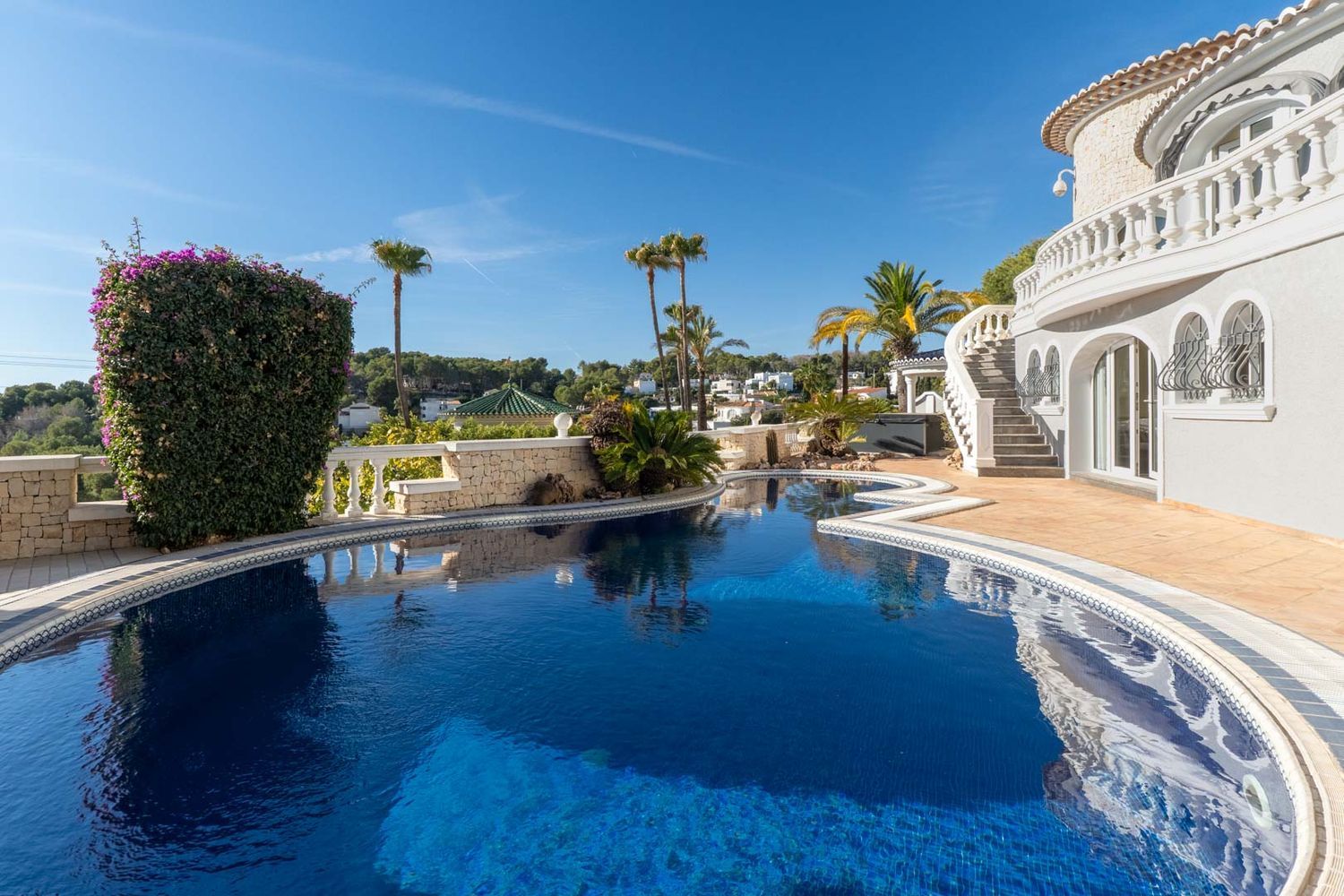 Großer, unregelmäßig geformter Privatpool mit blauen Mosaikfliesen, umgeben von hellen Pflastersteinen und mediterraner Bepflanzung. Eine geschwungene Treppe mit weißer Balustrade führt zur Villa.