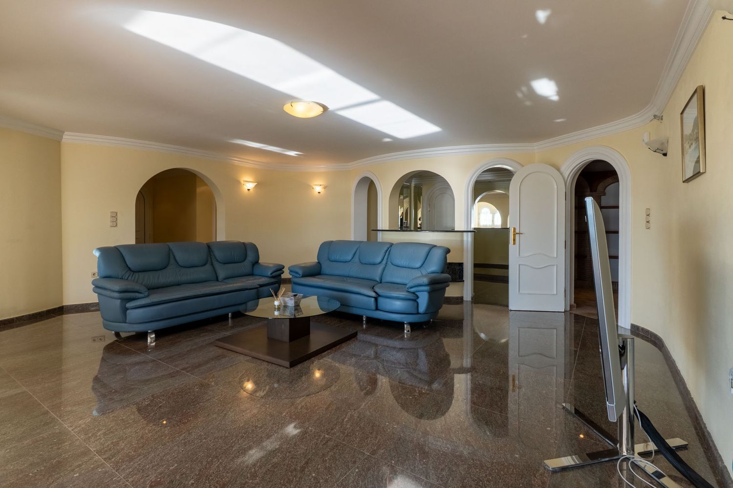 Großzügiger Wohnbereich mit dunklem, poliertem Marmorboden, blauen Ledersofas und mehreren Rundbögen zu angrenzenden Räumen. Natürliches Licht spiegelt sich an der Decke.