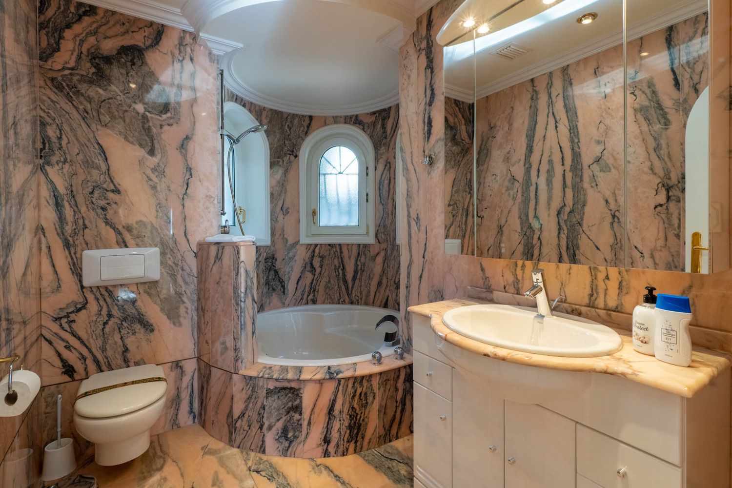 Hauptbadezimmer mit rosa-schwarz geädertem Marmor, einer eingebauten Badewanne in einer Rundnische und einem Bogenfenster.