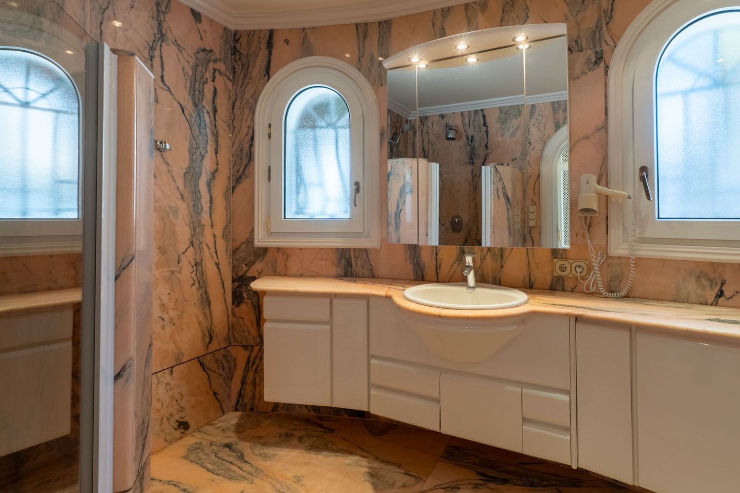 Hauptbadezimmer mit Wänden und Boden aus geädertem rosa Marmor, einem schwebenden weißen Waschtisch mit integriertem Waschbecken und zwei gewölbten Milchglasfenstern.