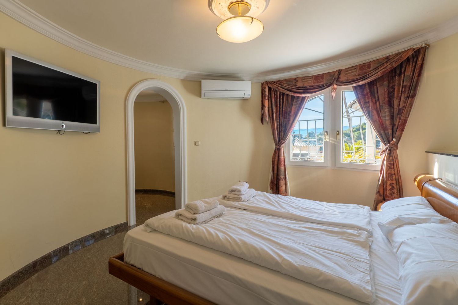 Helles Schlafzimmer mit zwei Einzelbetten, Wandfernseher und teilweisem Meerblick durch Fenster mit gemusterten Vorhängen. Klimaanlage und Zugang zu einem weiteren Raum.