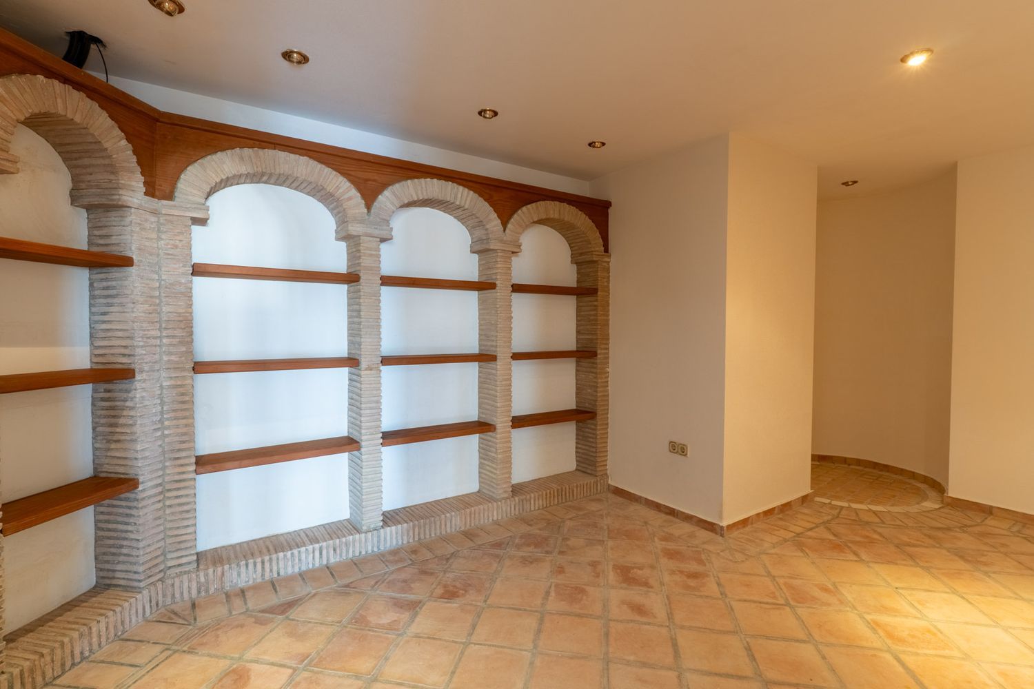 Innenraum mit Terrakotta-Bodenfliesen und gemauertem Regal mit Ziegelbögen und Holzbrettern. Einbaustrahler und eine markante Ecke mit rundem Bodendetail.