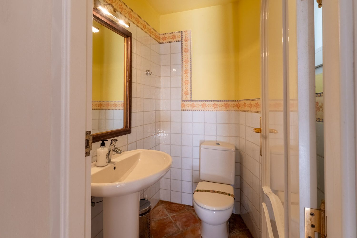 Kompaktes Badezimmer mit Standwaschbecken, Holzspiegel mit Oberlicht und weiß gefliesten Wänden mit floralen Zierleisten. Es umfasst eine Toilette und Terrakotta-Bodenfliesen.