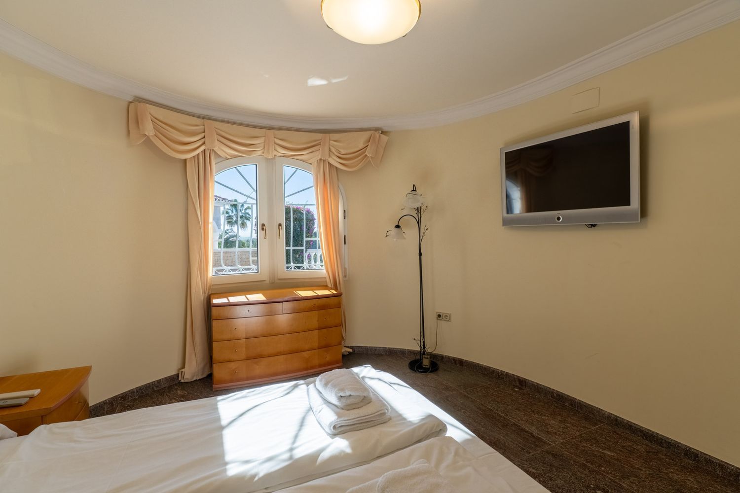 Schlafzimmer mit Bett, heller Holzkommode unter einem gewölbten Fenster und Wand-TV. Natürliches Licht erhellt den Raum mit dunklem Fliesenboden und Blick ins Grüne.
