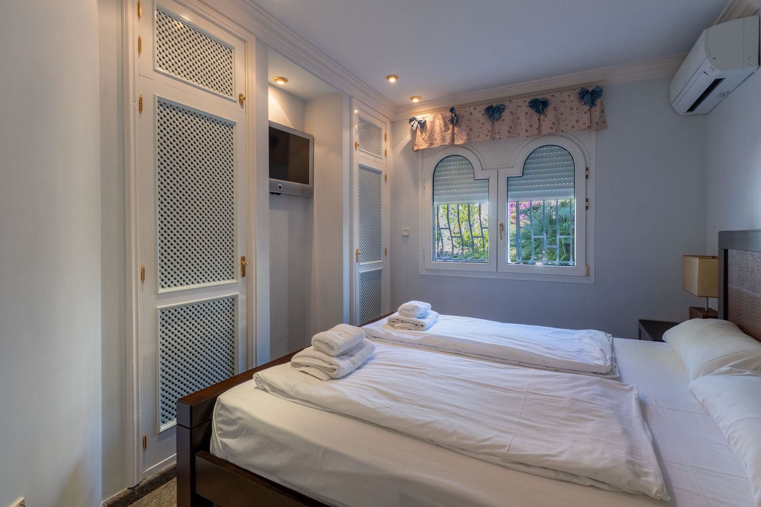 Schlafzimmer mit weißen Lamellen-Einbauschränken, Wandfernseher und gewölbten Fenstern mit Gartenblick.