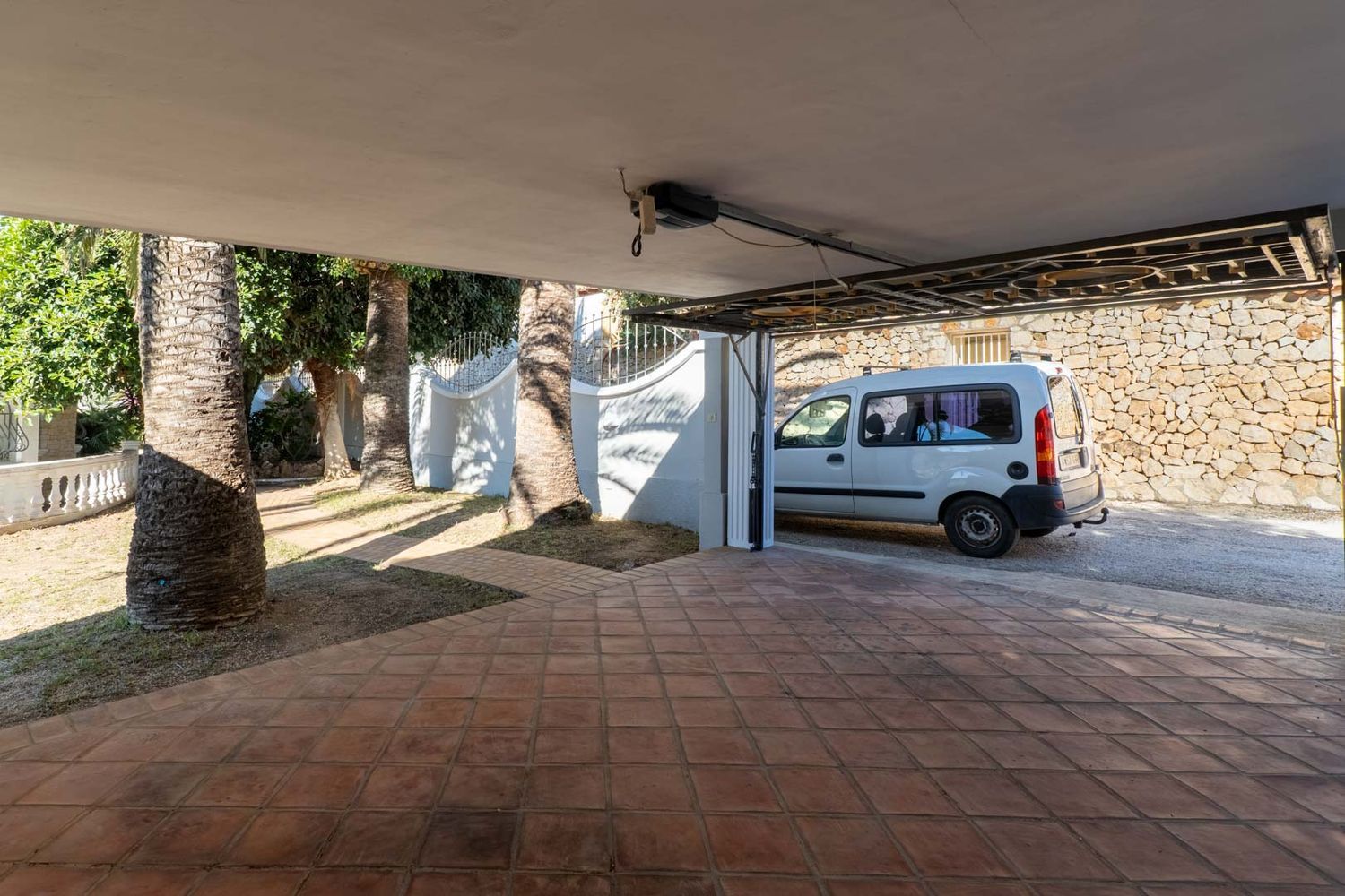 Überdachter Parkbereich mit Terrakottafliesen, umgeben von Palmen und einem geparkten weißen Lieferwagen. Eine Steinmauer ist im Hintergrund sichtbar.