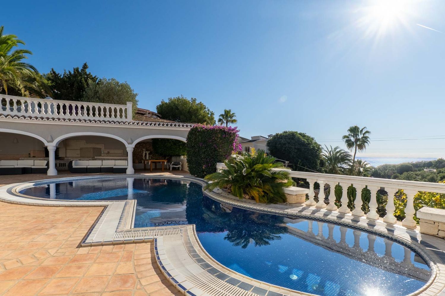 Unregelmäßig geformter blauer Mosaikpool mit Terrakottafliesen, angrenzend an eine überdachte Terrasse mit weißen Sofas. Fernblick auf das Meer und mediterrane Vegetation.