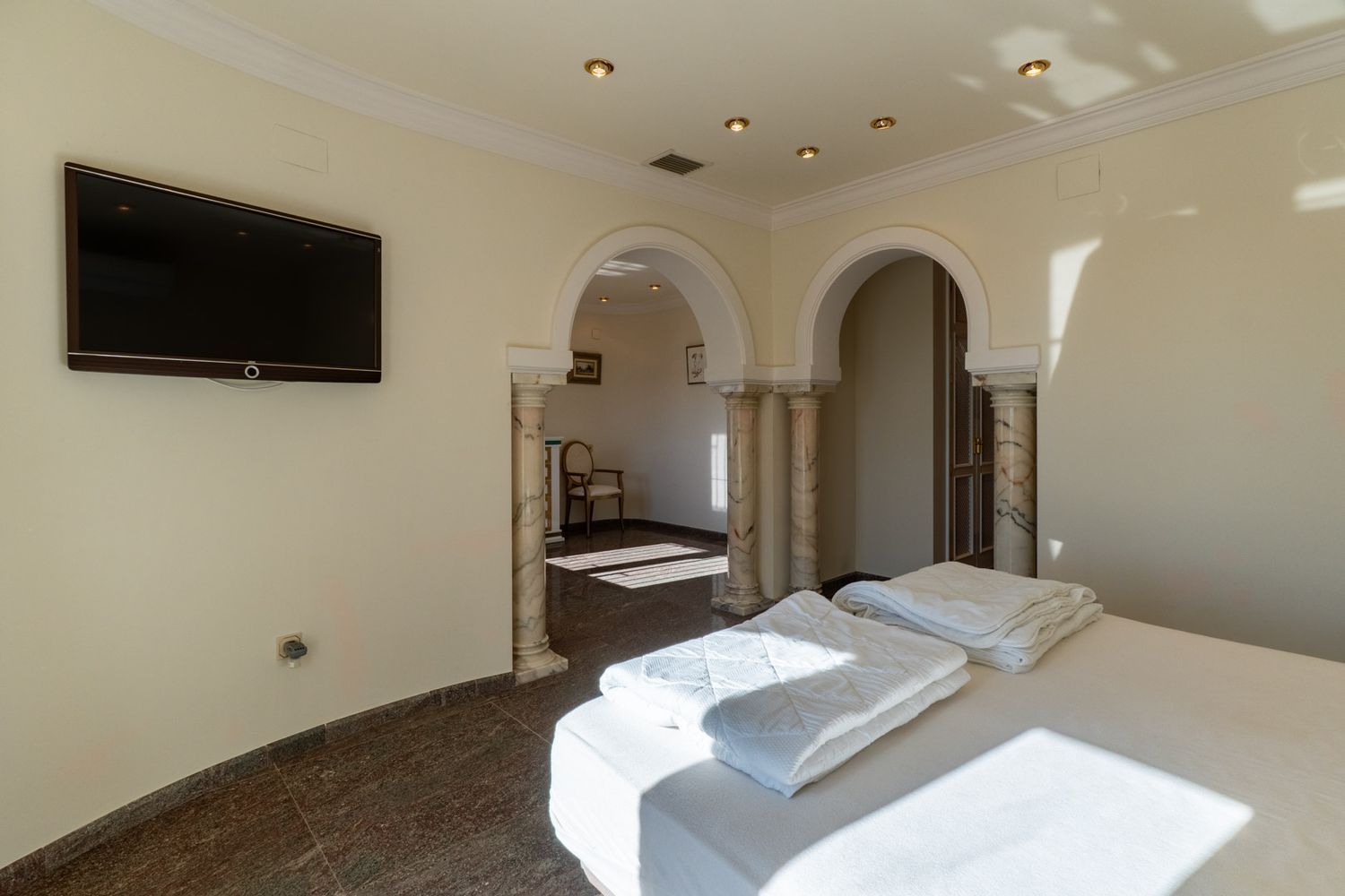 Zimmer mit Bett, dunklem Marmorboden und wandmontiertem Fernseher. Zwei gewölbte Durchgänge mit Marmorsäulen führen in einen Nebenraum, beleuchtet durch Einbauleuchten und natürliches Tageslicht.