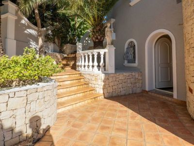 Außeneingang mit Terrakottafliesen und aufsteigender Treppe. Eine klassische weiße Balustrade und Natursteinmauern umrahmen die graue Fassade mit Bogentür.