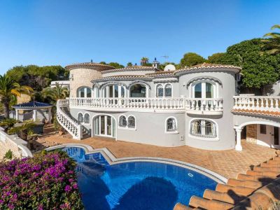 Die Villa zeigt eine graue Fassade, weiße Balustraden und einen markanten Privatpool. Eine geschwungene Treppe verbindet den oberen Balkon mit der terrakotta-gefliesten Poolterrasse, umgeben von mediterraner Flora.