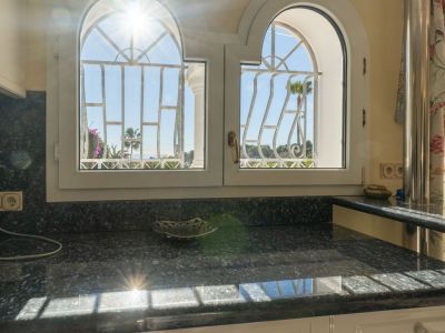 Dunkle Granitarbeitsplatte unter zwei Rundbogenfenstern mit Ziergittern, die teilweisen Meerblick und Palmen zeigen. Direktes Sonnenlicht erhellt die Oberfläche.