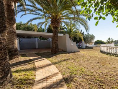 Ein gepflasterter Weg führt durch einen Garten mit Palmen und Rasen. Ein überdachter Carport und eine geschwungene weiße Balustrade sind sichtbar, umgeben von mediterraner Vegetation unter blauem Himmel.