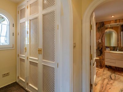 Flur mit weißen Einbauschränken mit Gittertüren und goldenen Griffen, neben einem Bogenfenster. Ein Rundbogen führt zu einem Badezimmer mit marmorierten Oberflächen.