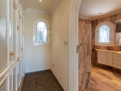 Flur mit weißen Lamellenschränken und goldenen Griffen, der zu einem Badezimmer mit rosa Marmorverkleidung und zwei Rundbogenfenstern führt.