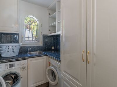 Funktionale Waschküche mit dunkler Granitarbeitsplatte, Spüle und Waschmaschinen. Das Rundbogenfenster mit Gitter sorgt für natürliches Licht, ergänzt durch weiße Schränke.