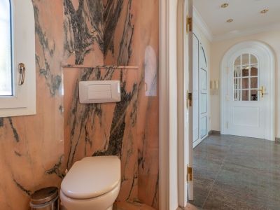 Gäste-WC mit rosa geäderten Marmorwänden und Fenster, das zu einem Flur mit dunklen Fliesen und einer Bogentür führt.