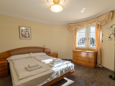 Geräumiges Schlafzimmer mit Doppelbett, Holz-Kopfteil und passenden Nachttischen. Natürliches Licht fällt durch ein geschwungenes Fenster auf den dunklen Fliesenboden.
