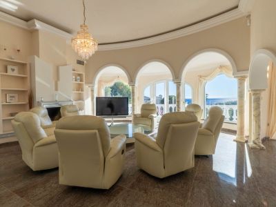 Geschwungenes Wohnzimmer mit Relaxsesseln, poliertem Marmorboden und Meerblick durch Bogenfenster. Ein zentraler Kristallleuchter und klassische Säulen prägen den eleganten Raum.