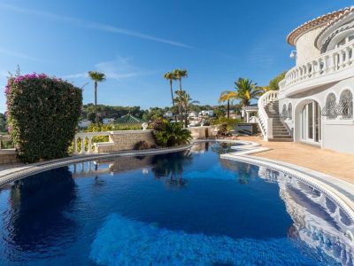Großer, unregelmäßig geformter Privatpool mit blauen Mosaikfliesen, umgeben von hellen Pflastersteinen und mediterraner Bepflanzung. Eine geschwungene Treppe mit weißer Balustrade führt zur Villa.