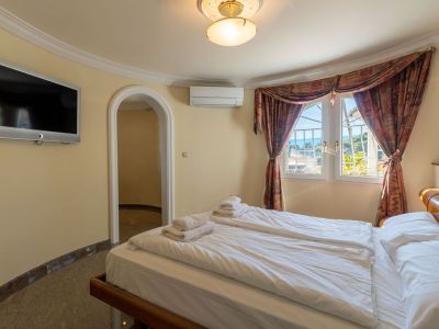 Helles Schlafzimmer mit zwei Einzelbetten, Wandfernseher und teilweisem Meerblick durch Fenster mit gemusterten Vorhängen. Klimaanlage und Zugang zu einem weiteren Raum.