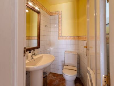 Kompaktes Badezimmer mit Standwaschbecken, Holzspiegel mit Oberlicht und weiß gefliesten Wänden mit floralen Zierleisten. Es umfasst eine Toilette und Terrakotta-Bodenfliesen.