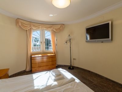 Schlafzimmer mit Bett, heller Holzkommode unter einem gewölbten Fenster und Wand-TV. Natürliches Licht erhellt den Raum mit dunklem Fliesenboden und Blick ins Grüne.