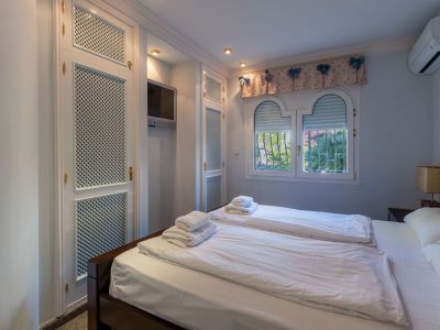 Schlafzimmer mit weißen Lamellen-Einbauschränken, Wandfernseher und gewölbten Fenstern mit Gartenblick.