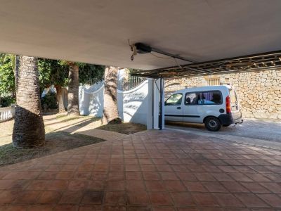 Überdachter Parkbereich mit Terrakottafliesen, umgeben von Palmen und einem geparkten weißen Lieferwagen. Eine Steinmauer ist im Hintergrund sichtbar.