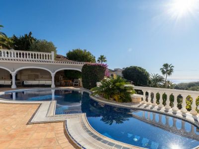 Unregelmäßig geformter blauer Mosaikpool mit Terrakottafliesen, angrenzend an eine überdachte Terrasse mit weißen Sofas. Fernblick auf das Meer und mediterrane Vegetation.