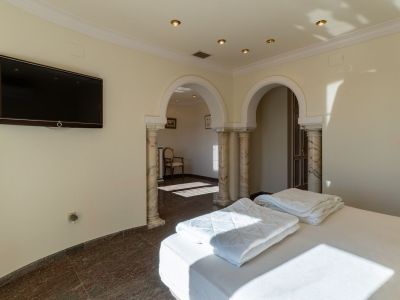 Zimmer mit Bett, dunklem Marmorboden und wandmontiertem Fernseher. Zwei gewölbte Durchgänge mit Marmorsäulen führen in einen Nebenraum, beleuchtet durch Einbauleuchten und natürliches Tageslicht.