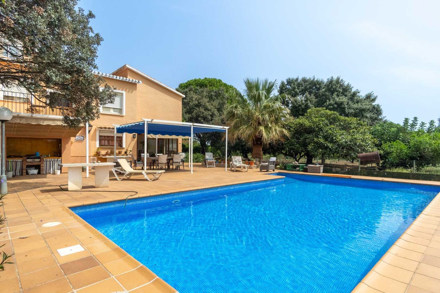 Großer Pool mit blauen Mosaikfliesen und gefliester Sonnenterrasse. Daneben ein schattiger Essbereich und eine Außenküche.