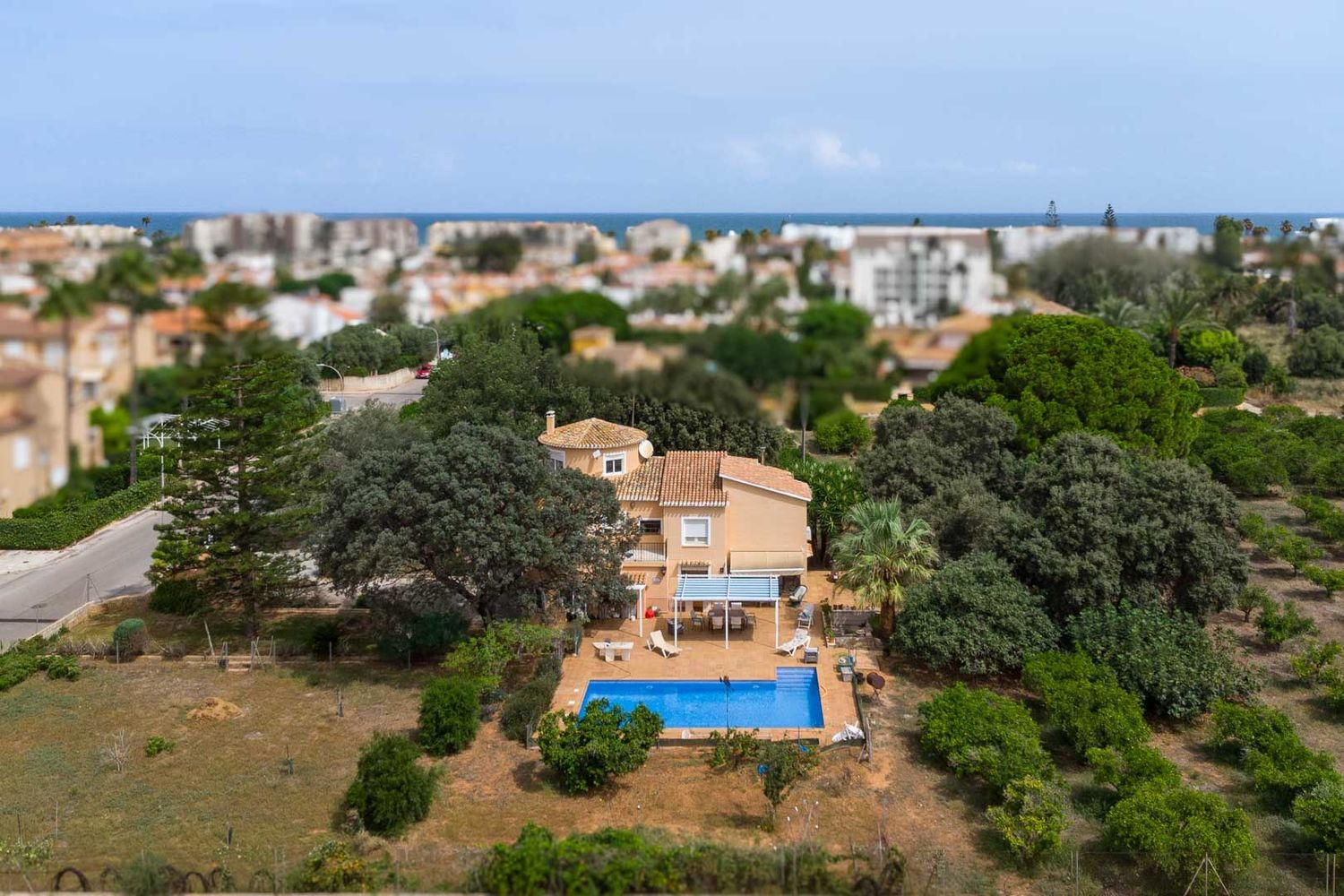 Luftaufnahme der Villa mit blauem Pool, schattiger Terrasse und großem, bewachsenem Garten. Im Hintergrund sind die Stadtkulisse und das Mittelmeer erkennbar.